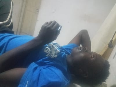 A person is leaning on their left hand and looking downwards, wearing a blue t-shirt with a white logo. A wall is behind them. The person is likely in an indoor setting. The image is slightly tilted. They appear to be wearing athletic shorts with white stripes down the side.
