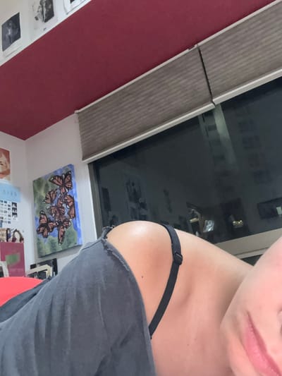 A photograph shows the partial view of a person from the shoulders up, the person is wearing a gray top with a black bra strap visible. The background features a window with blinds partially obscuring the view, which appears to be an urban setting at night. Below the window, on the wall, is a painting of butterflies and a collection of smaller pictures or prints. The location is potentially Jesús del Monte, Mexico.