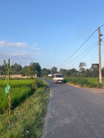 The image depicts a scene on a rural road in Lendangnangka Utara, Indonesia, likely during the late afternoon given the warm lighting and long shadows. The perspective is from a low angle, looking upwards towards a bright blue sky with scattered wispy clouds. A tall utility pole with numerous wires dominates the left side of the frame, extending out of view at the top.

The road itself, paved with asphalt, curves gently to the right and is bordered by lush green vegetation. On the left side of the road, there are rice paddies, their vibrant green color contrasting with the dusty grey of the road. Tall grasses and trees form a dense natural barrier along the roadside.

A white van is the only vehicle visible, traveling along the road away from the viewer. The van has a license plate that appears to read "BP 8723 SE." Attached to the utility pole is a dark rectangular sign with white text, which reads "Ombak Hidden Villa." This suggests the location is near a villa or resort.

In the foreground, a plant with long, strap-like leaves is visible, with a crumpled green plastic bag tied to its stem. The overall impression is of a peaceful, tropical setting, characteristic of rural Indonesia. The weather appears clear and sunny.