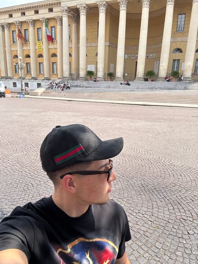 A medium shot of a man wearing a black Gucci baseball cap and black Ray-Ban sunglasses in the city of Verona, Italy. The man wears a black t-shirt with a colorful design. The paving stones of the Piazza Bra are visible. In the background, the Palazzo della Gran Guardia is also visible. People are sitting on the steps of the building.