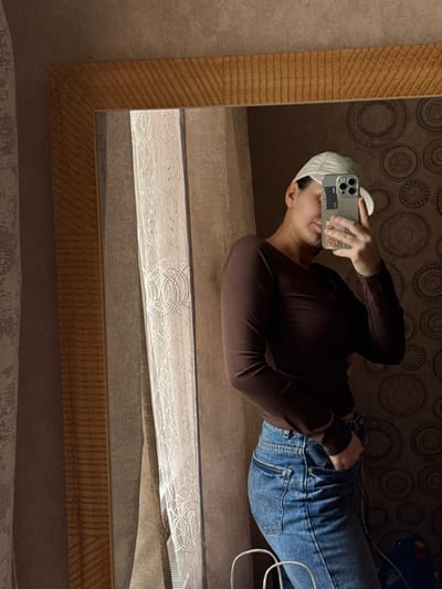 The image depicts a female individual taking a mirror selfie within an indoor residential space in Velikovechnoye, Russia. The subject is partially visible from the chest to the upper thighs, wearing a dark brown long-sleeved shirt, blue denim jeans, and a light-colored baseball cap. She holds a silver-toned smartphone with a patterned case in her left hand, obscuring her face, while her right hand is placed in the right front pocket of her jeans. The reflection is captured in a rectangular mirror framed by textured, light brown wood. The background consists of a wall with two distinct wallpaper patterns: vertical stripes in shades of brown and tan to the left, and a circular, possibly mosaic-style pattern in brown tones to the right. Light-colored, translucent curtains are visible to the left, suggesting a window. A white wire object is partially visible in the lower foreground, and a dark object with blue and yellow elements is discernible in the lower right background, partly obscured by the subject's reflection.