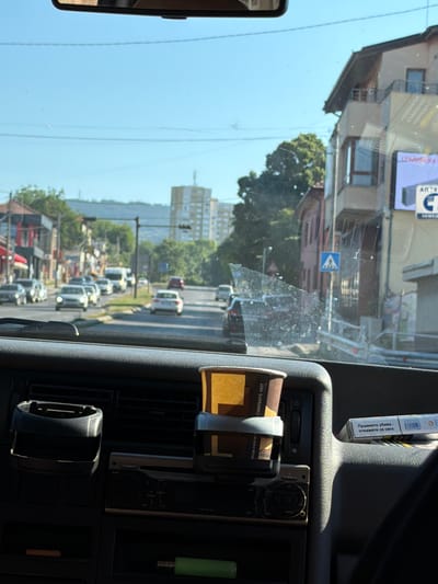 This image, taken from inside a vehicle on a sunny day, offers a glimpse of a street scene in Radomir, Bulgaria. The foreground is dominated by the dashboard of the vehicle, featuring a cup holder with a disposable coffee cup, a car stereo, and a pack of cigarettes. The windshield reveals a street with moderate traffic, including cars and a white van. Buildings line the street, some appearing residential with balconies, while others are commercial. In the background, taller apartment buildings and trees are visible against a clear blue sky. A pedestrian crossing sign is noticeable on the right side of the street. The overall impression is that of a typical urban environment in Bulgaria, likely captured during daylight hours.