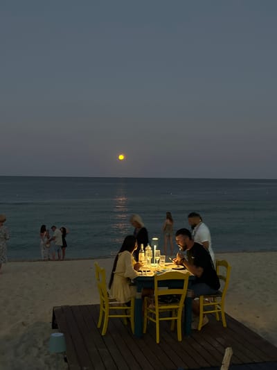 The image depicts an outdoor dining scene on a sandy beach at dusk, with the sea and a large, bright moon visible in the background. A group of people are gathered around a table set for dinner. The table is lit by a lamp, illuminating the food and drinks. One man is actively eating, while others appear to be engaged in conversation. In the middle ground, several other individuals are standing near the water's edge, some seemingly interacting with each other. The overall atmosphere suggests a relaxed evening gathering by the sea. The scene appears to be set on a beach in the Toroni Municipal Unit, Greece, given the presence of the sea and the characteristic natural landscape.