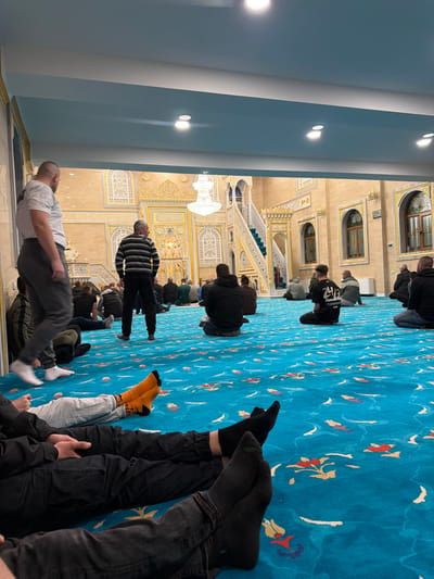 The image depicts the interior of a mosque in Breznitsa, Bulgaria, observed from a low-angle perspective. The floor is covered with a vibrant blue carpet featuring abstract floral patterns in shades of red, yellow, and white. In the foreground, the lower bodies of two individuals are visible, seated on the carpet with their legs extended; one wears dark pants and black socks, the other grey sweatpants and bright orange socks.
Approximately 20-30 adult males are present within the large hall. Most are seated on the carpet, facing towards the ornate back wall, some cross-legged, others with legs extended. A man in a white t-shirt and grey pants walks barefoot, wearing white socks, from left to right in the middle ground. Another man in a black and white horizontal striped long-sleeve shirt stands facing the back of the room.
The architectural elements include beige/light brown walls extensively decorated with intricate white and gold carvings, arches, and panels. A large, multi-tiered crystal chandelier hangs centrally from the ceiling. A white and gold minbar with green steps is visible on the right side of the background. Arched wooden-framed windows are present on the right wall. The ceiling is light blue with white trim and multiple recessed lights.