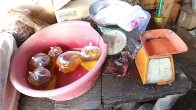 An overhead, medium shot shows a pink plastic basin filled with several small, clear plastic bags containing a yellow liquid, tied with red and white strings, set on a dark, dirty wooden surface. To the right of the basin is an orange and white scale with an empty orange tray. To the left and behind the basin are cardboard boxes, one with "65590059 LIME" printed on it. A green and yellow handled object, possibly a broom or mop, stands behind the cardboard boxes. The scene appears to be an outdoor market stall in Puloara, Indonesia.