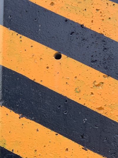The image is a close-up, vertical shot of a concrete surface painted with diagonal black and yellow hazard stripes. The concrete has a rough texture with numerous small pores and some larger pits, suggesting it is an exterior surface. There is a small, dark circular hole visible in the upper central portion of the image, on a yellow stripe. The paint appears to be slightly worn in places, with some areas showing peeling or chipping, particularly on the yellow stripes. The lighting is even, and there are no discernible shadows, indicating an overcast sky or diffuse lighting conditions. The image does not depict any people, actions, or interactions. It is a detail shot of a functional, safety-oriented surface. The presence of hazard stripes suggests a location such as a construction site, a parking garage, a loading dock, or a barrier in a public area. Without further context or identifying markers, the specific location within Chiang Rai City Municipality, Thailand, cannot be determined from the visual information alone.