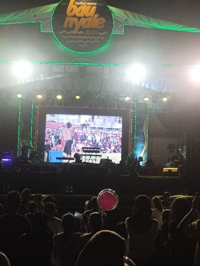 This night photograph captures an outdoor festival or concert in Kecamatan Pujut, Indonesia, likely the "Festival Pesona Bau Nyale 2025," as indicated by the prominent green neon-lit circular sign above the stage. A large crowd of attendees, visible primarily as dark silhouettes, faces the brightly illuminated stage.
The stage features a large central LED screen displaying a performance, possibly a live feed or a pre-recorded segment, showing a person in traditional attire engaging with a smaller crowd. The stage is equipped with numerous bright spotlights and truss structures, casting a strong glow and illuminating the screen. Silhouettes of crew members or performers are visible on the stage floor, along with what appears to be musical equipment, including a keyboard stand and possibly a guitar.
In the foreground, a distinct red or pink balloon-like object, seemingly illuminated, stands out among the audience. The overall setting is dark, emphasizing the bright stage lights and screen, confirming it is nighttime. The crowd appears engaged, watching the event unfold on the stage and screen.