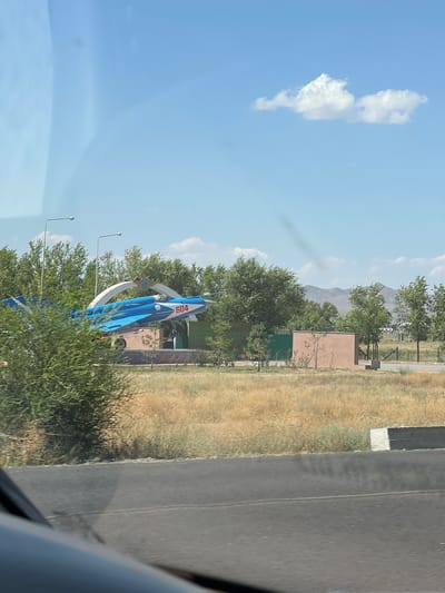 An outdoor scene in Saro-bulak, Kazakhstan, captures a bright, clear day with a few wispy clouds in the pale blue sky. The foreground shows a portion of a dark asphalt road, possibly with a yellow line. Beyond the road, dry, golden-brown grass covers the ground. In the mid-ground, a monument featuring a blue and white fighter jet with the number "604" visible on its side stands prominently. The jet is mounted on a concrete pedestal and is surrounded by greenery, including trees and bushes. Two tall lampposts are positioned behind the jet. To the right of the jet monument, there is a small, light brown rectangular building and a green fence. In the background, rolling hills are visible under the vast sky, and the landscape appears somewhat arid. The image seems to be taken from a moving vehicle, as indicated by the slight blur and the reflection of something on the windshield in the upper left corner.
