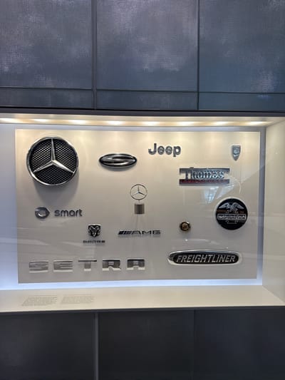 This media file captures a display of automotive brand emblems, predominantly from Mercedes-Benz and its associated brands. The objects are mounted on a plain, light-colored background within a display case. To the left and right of the display are textured, dark gray panels, suggesting a modern architectural setting.
The subjects are a collection of chrome and metallic emblems. Visible brands include "Jeep," "Thomas Built Buses," "American LaFrance," "Freightliner," "Mercedes-Benz" (represented by the star emblem), "AMG," "Smart," and "Dodge." There are also individual letters arranged vertically that spell out "SETRA," another bus manufacturer. A circular emblem with wings and text beneath it, likely "American LaFrance," is also present. A small, circular emblem with a ram's head is visible, which is the Dodge logo. A three-pointed star emblem is prominently displayed, as is a circular mesh emblem with a star in the center.
The scene is a static display, with no people or ongoing activity evident. The lighting appears to be artificial and is integrated into the display case, casting subtle shadows on the wall behind the emblems. There are no obvious cues regarding the time of day or weather. The location context of Stuttgart, Germany, suggests this might be a museum exhibit or a dealership display showcasing the history and brands associated with the automotive industry in that region.
The text visible includes brand names such as "Jeep," "Thomas Built Buses," "American LaFrance," "Freightliner," "AMG," "Smart," "Dodge," and "SETRA." There is also some small, unreadable text on a white strip on the right side of the display, possibly an informational plaque. The overall impression is one of a curated collection of automotive heritage.