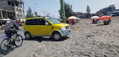 The image depicts a yellow minivan parked on the side of a paved road in Rubavu, Rwanda, located at latitude -1.668 and longitude 29.269. The minivan is facing the right side of the image, with its front wheels slightly raised off the ground. A man on a blue bicycle is traveling in the opposite direction of the minivan, riding on the left side of the road. The background features a multi-story building and several trees. The road is paved with uneven, gray stone blocks. There is a large, red, open umbrella in the background, on the right side of the image, with two individuals sitting under it. The ground is dry and dusty. Several other people are visible in the distance, walking on the road and sidewalk. The sky is clear and blue.