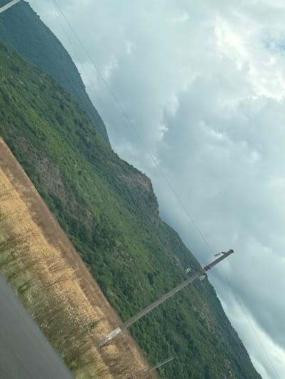The image displays a diagonal view of a mountainous landscape in Ismailli District, Azerbaijan. A steep, green, tree-covered mountain dominates the right side of the frame, rising towards a cloudy sky. In the foreground, a strip of dry, yellowish-brown grass with scattered white flowers runs diagonally from the bottom left, separating the mountain from a dark asphalt road on the far left. A utility pole with insulators and power lines stretches vertically across the mid-right of the image, positioned in front of the mountain. The sky is overcast with large, soft gray clouds. The scene appears to be an outdoor rural or semi-rural environment.