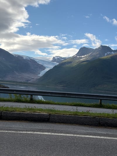 The image displays a scenic view of a glacial valley in Svartisen Gård, Norway. In the background, a large glacier flows down a mountainside, feeding into a calm body of water that reflects the surrounding landscape and the sky. The mountains are covered in lush green vegetation on their lower slopes, with patches of snow visible higher up. A grey metal guardrail runs horizontally across the mid-ground, separating the roadside from the water. The foreground shows an asphalt road with a white line, bordered by a stone curb and a strip of green grass with some small wildflowers. The sky is partly cloudy, with patches of blue sky visible between fluffy white clouds. The overall impression is one of natural grandeur and serene beauty.
