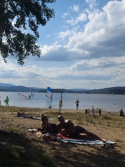 The image depicts a sunny day at a lake, likely the Radslav Reservoir in the Czech Republic, based on the provided context of Radslav. In the foreground, two individuals are relaxing on a beach towel, positioned on the sandy shore. One person, with fair hair styled in a bun and wearing sunglasses, is lying down on their side, facing towards the left. Next to them, a person wearing black swim trunks is lying on their back, also facing towards the left. In the middle ground, several people are engaged in water sports. Two individuals are windsurfing on the lake, one with a blue and white sail and the other with a red sail that appears to have fallen. Further out on the water, several sailboats are visible, and a person is standing on a paddleboard. The background features a tree line with forested hills and a bright blue sky with scattered clouds. The beach is sandy with patches of dry grass.
