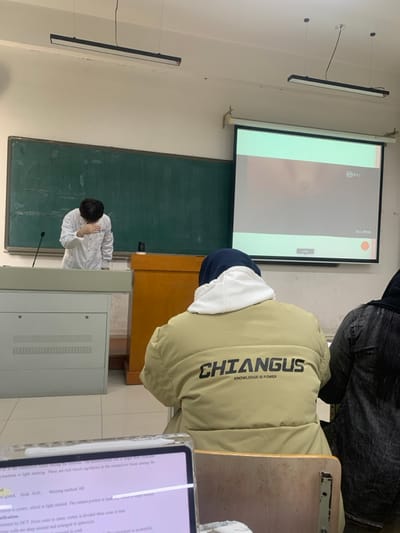 A lecture hall scene with a student at the podium. A presentation is displayed on a screen, possibly a video call or lecture recording. Students are seated, one wearing a tan jacket with "CHIANGUS" printed on the back. An iPad rests on a desk, displaying text likely related to the lecture. A chalkboard is visible on the wall, suggesting a traditional classroom setting integrated with modern technology.