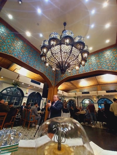 A lavishly decorated restaurant interior is captured in an angled, well-lit shot. A large, ornate chandelier with intricate patterns dominates the upper left, complemented by patterned wall decor. Tables are occupied by people; some are seated while one man in a suit seems to be taking a photo. A prominent, oversized glass orb, likely a decorative light fixture, occupies the lower right, slightly obscuring the scene. Large arched windows line the rear of the space.