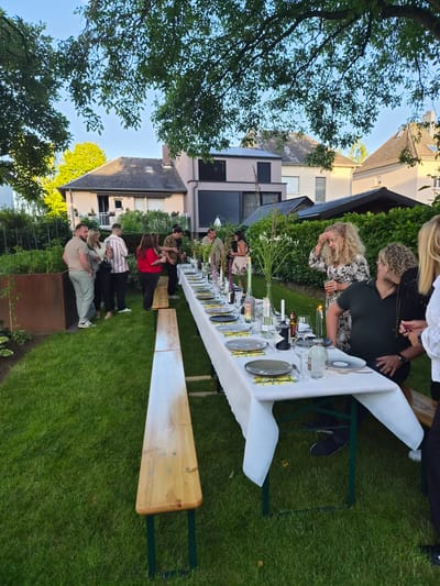 In Bettembourg, Luxembourg, a group of approximately ten adults are gathered in a lush green garden for an outdoor dinner party. A long table, covered with a white tablecloth, is set with plates, glasses, and bottles of drinks, indicating a meal is in progress. People are standing and seated around the table, engaged in conversation. The setting appears to be a residential backyard, with houses visible in the background and mature trees providing shade. The lighting suggests it is late afternoon or early evening.
