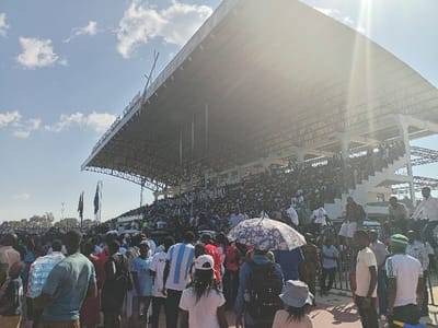 The image depicts a large crowd gathered at an outdoor stadium under a bright, sunny sky in Siaya, Kenya. The stadium features a prominent, cantilevered roof structure covering multiple rows of spectator seating, which are densely packed with attendees. Visible on the facade of the stadium's upper structure, partially obscured by sun glare, is the text "SIAYA STADIUM."
In the foreground, a diverse crowd of people is observed from behind, standing and moving within the stadium grounds. Individuals are dressed in varied casual attire; notable details include a person wearing an Argentina national football team jersey, another holding a patterned umbrella for shade, and several carrying backpacks. The scene suggests a significant public event, such as a sporting match, political rally, or large community gathering.
The time of day appears to be midday, indicated by the high sun causing a bright lens flare in the upper right and casting strong illumination across the scene. The weather is clear with a few scattered white clouds against a blue sky. Flags are visible on poles to the left, beneath the stadium roof. The overall atmosphere is bustling with a large number of people present.