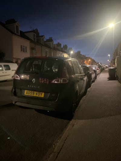 The image depicts a dark street scene in East Suffolk, United Kingdom at night. Cars are parked along one side of the street. A black Renault Scenic with the license plate A059 VOY is prominently featured in the foreground, positioned close to the curb. Buildings, likely residential, line the street on the opposite side of the parked cars. Streetlights illuminate the scene, casting light and shadows.