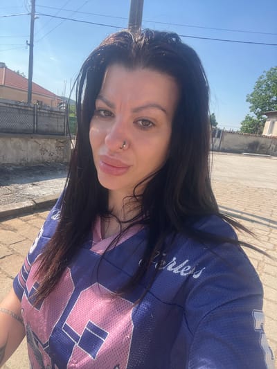 In Zelenikovo, Bulgaria, a young woman with dark hair and a nose ring takes a selfie outdoors. She is wearing a blue and pink jersey. The background shows a street paved with cobblestones, a building with a red-tiled roof, a fence, and trees under a clear blue sky.