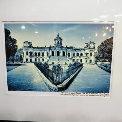 The image displays a physical print of the Tajhat Zamindar Palace in Rangpur, Bangladesh. The photograph itself exhibits a strong blueish, almost monochromatic, tone, lending it a historic or stylized aesthetic. The central subject is the grand, symmetrical palace, characterized by a prominent central dome, flanked by two side towers, numerous arched windows, balconies, and intricate architectural embellishments. A broad, multi-tiered staircase leads up to the main entrance of the building.
In the foreground, a paved pathway guides the eye towards the palace. This pathway is bordered by a V-shaped garden plot, meticulously arranged with small, manicured bushes, and enclosed by a dark metal fence. Large, leafy trees frame both sides of the palace in the background. The sky above is bright with scattered clouds, indicating that the scene was captured during daytime. Notably, several reflections from overhead light sources are visible on the surface of the print, suggesting it is displayed behind glass or a protective covering.
No people or discernible activities are present in the image, allowing the focus to remain on the architectural grandeur of the palace.
The text visible beneath the image provides contextual information: "Tajhat zamindar palace, Rangpur From 1984 to 1991 Tajhat palace, Rangpur was used as the High Court bench of the Supreme Court of Bangladesh". Although the provided location context for the media file is Bhola, Bangladesh, the subject of the photograph is specifically identified as being located in Rangpur.