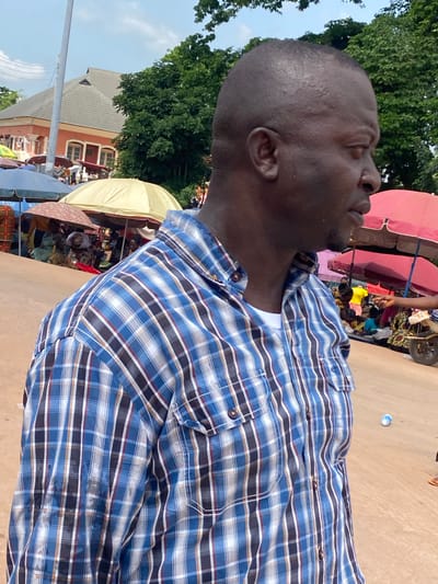 In Igbara, Nigeria, a medium shot captures a man from the chest up, positioned in profile and facing right. He is wearing a blue and white checkered long-sleeved shirt over a white undershirt. His head is bald, and he appears to be at an outdoor market. In the background, several stalls with umbrellas are visible, and people are gathered around them. Trees and a building with a red facade and a gray roof are also in the background. The ground is a light brown dirt path.
