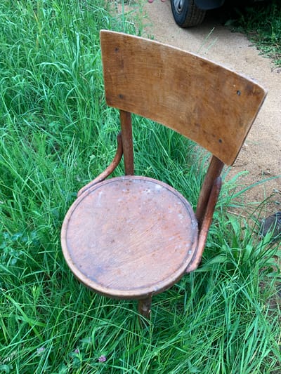 A weathered, wooden chair with a curved back and round seat sits on lush green grass and a dirt path. The chair appears to be old and shows signs of wear, with water stains and visible nail heads. The background features tall, dense grass and a sandy or gravelly path, suggesting an outdoor setting possibly in a rural or less-maintained area. A small portion of a car tire and fender are visible in the upper right corner, partially obscured by the chair and grass. The lighting suggests it is daytime, likely overcast given the lack of harsh shadows. The scene is devoid of human presence, focusing entirely on the static object within its natural environment. The location, Блеўчыцкі сельскі Савет, Belarus, indicates a village or rural administrative region.