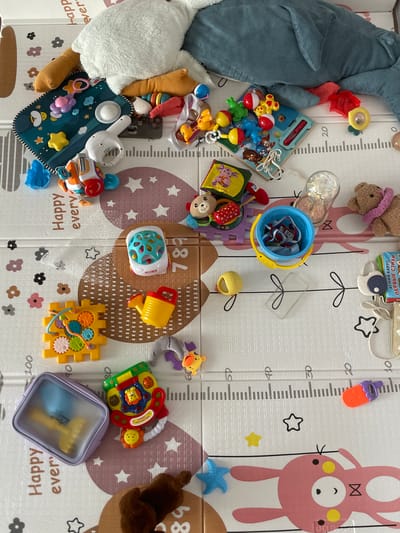 The image depicts a collection of baby toys scattered across a play mat. The play mat features a grid design with height measurements along the edges, along with repeating "Happy every" text and illustrations of stars and flowers.
The toys include a stuffed shark, a plush bear, a blue bucket with items inside, a musical toy, a set of gears, a plush giraffe, and other various play items. The toys are spread out across the mat, indicating a child's play area.
The OCR text on one of the toy packages is identified as Russian "АГНИЯ БАРТО ПЕРВЫЕ СТИХИ" (Agniya Barto First Poems). The mat also has "СХБ" as OCR text.
There is no indication of the location being in the city of Средний Постол, Russia. There are no people in the image.