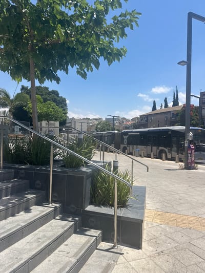 The image depicts an outdoor urban scene in Bnei Brak, Israel, on a sunny day. In the foreground, a set of grey stone stairs with metal railings leads upwards. Adjacent to the stairs is a planter box filled with lush green foliage.
The midground features a row of modern buses parked along a paved area. The buses are predominantly grey with dark windows and some display logos and text in Hebrew. A street light fixture is visible on the right side of the image, along with several bollards. A trash bin with Hebrew writing is also present near the buses.
The background reveals a city skyline with a mix of buildings, including multi-story residential structures and some taller commercial buildings. Trees with green foliage are scattered throughout the urban landscape, and the sky is a clear blue with a few scattered clouds. The overall impression is a functional urban transportation hub.