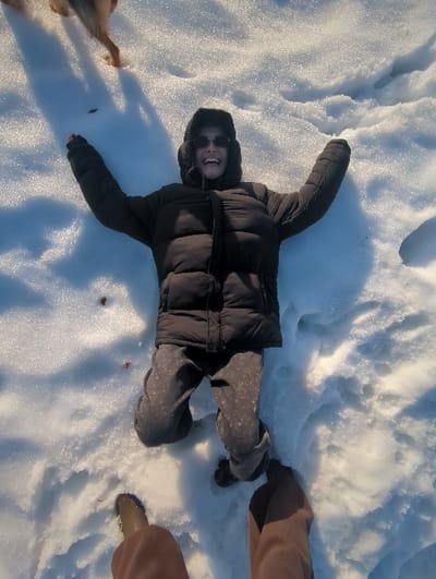 An overhead view depicts a person lying on their back in white snow, surrounded by foot impressions and areas of disturbed snow. The individual, positioned centrally, is wearing a dark, possibly black or very dark brown, puffy winter jacket, grey sweatpants with a speckled pattern, and dark shoes. They have a hood up, earflaps visible, and are wearing dark sunglasses, smiling broadly with an open mouth. Their arms are outstretched to the sides, hands near their head. In the upper left background, the hindquarters and tail of a light-brown dog are partially visible, along with its elongated shadow cast on the snow. In the immediate foreground, the lower legs and feet of another individual, presumably the photographer, are visible; one foot wears a light brown boot, and the other is obscured by brown pants. A few small, reddish-brown specks are scattered on the snow near the central figure's left arm. Strong shadows on the snow indicate bright sunlight. The scene is located in Podolino, Russia.