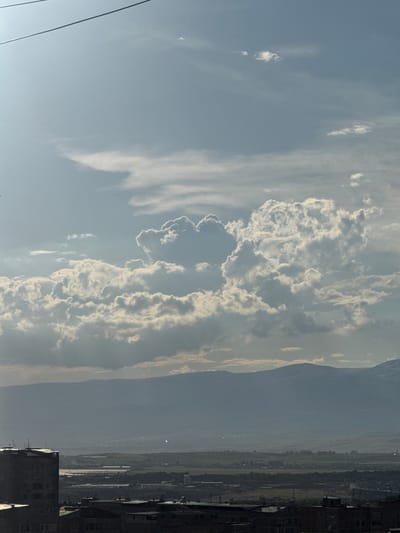 This is a wide shot of a cityscape with mountains in the background, taken from a high vantage point, possibly a rooftop or an elevated window. The sky is a pale blue, with large, fluffy white clouds scattered across it. In the foreground, there are several buildings, some with flat roofs and others with sloped roofs. A tall, dark building with many windows is prominent on the left side of the image. In the distance, a range of hazy mountains forms the horizon. The middle ground shows a sprawling urban area with smaller buildings and what appears to be agricultural land. The overall impression is of a sunny day in a city located in a mountainous region, likely Abovyan, Armenia, given the context.