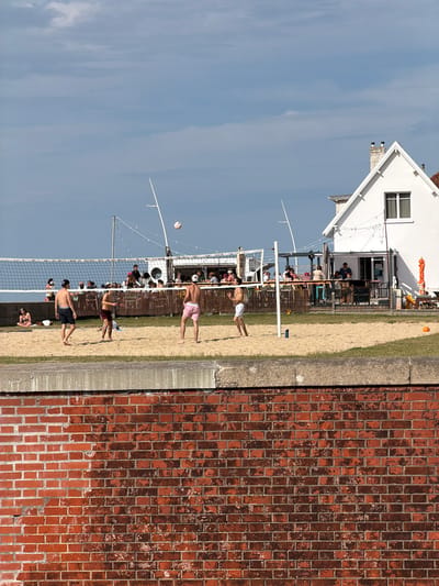 The image depicts an outdoor scene on a sunny day, likely in the afternoon given the bright light and shadows. The primary subjects are people playing beach volleyball on a sandy court. Several individuals are visible in various stages of the game, including jumping, diving, and serving. A volleyball is in mid-air, indicating an active play.

The setting is a beach volleyball court with a net stretched across it. In the background, there is a white building with a distinctive roofline, possibly a restaurant or clubhouse, with people seated at tables under an awning. Beyond the building, the sky is a clear blue with a few wispy clouds. To the right of the frame, a weathered red brick wall runs vertically, suggesting a boundary or part of a structure. A concrete ledge is visible at the base of the brick wall.

The mood appears to be relaxed and recreational, with people enjoying a casual game in a pleasant environment. The weather is clearly sunny and warm, ideal for outdoor activities. There are no visible text elements in the image. The overall scene suggests a holiday or leisure setting, common in coastal areas. The presence of the beach volleyball court and the building with outdoor seating points towards a public recreational area or a beach resort.