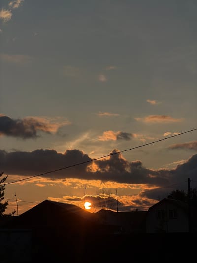 The image depicts a sunset over a residential area in Emel'yanovo, Russia. The sun, a bright orb of orange light, is partially obscured by dark, dramatic clouds, casting warm hues across the sky. Silhouetted against the sunset are the dark roofs of several houses, with the sharp peak of one prominent roof in the foreground. Electrical wires stretch horizontally across the frame, dividing the sky and adding a man-made element to the natural scene. The sky itself is a gradient of soft blues and oranges, with lighter clouds catching the last rays of sunlight. The overall impression is one of a peaceful, albeit dramatic, end to the day in a rural or suburban setting.