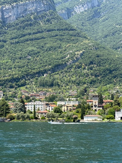 A wide shot captures a sun-drenched scene of Tremezzina, Italy, with the deep blue waters of Lake Como in the foreground. The lake is active with a white speedboat creating a foamy wake as it speeds across the water, carrying two visible passengers. Behind the lake, a dense green hillside rises sharply, covered in lush trees and dotted with a scattering of charming houses and villas. The architecture of the buildings is varied, with some featuring red-tiled roofs and others painted in lighter colors. Several olive trees are visible, adding to the Mediterranean feel of the landscape. In the distance, a massive, sheer rock face dominates the upper portion of the frame, partially shrouded in shadow. The overall impression is one of natural beauty and serene lakeside living.