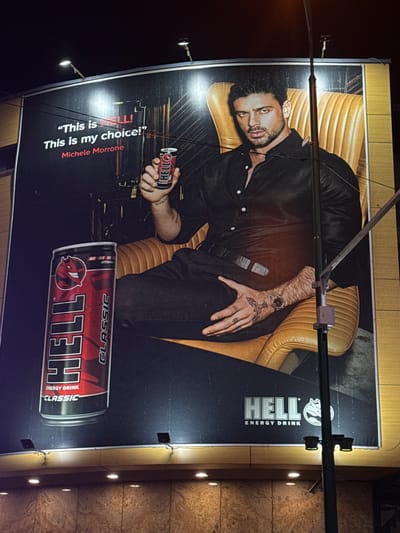 This is a nighttime, exterior, medium-shot photograph of a large billboard in Ilidža, Bosnia and Herzegovina, promoting "Hell Energy Drink." The billboard features a man, identified as Michele Morrone, seated in a golden-brown armchair. He is wearing a black collared shirt, dark pants, and a belt. Morrone holds a red can of "Hell Energy Drink" in his right hand and is looking directly at the camera. The text "This is HELL! This is my choice! Michele Morrone" is displayed prominently on the billboard. To the left of the man, a large, red can of "Hell Energy Drink Classic" is depicted vertically. The "Hell Energy Drink" logo is also visible in the lower right corner of the billboard. The billboard is illuminated by external lights. The surrounding environment suggests an urban setting at night, with visible streetlights and building elements at the bottom of the image.