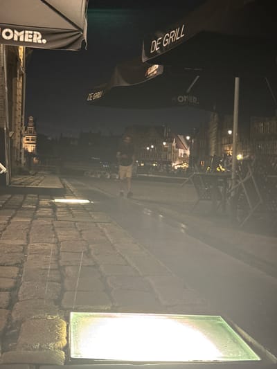 This is a night-time photograph taken outdoors on a cobblestone street in Ghent, Belgium, likely in a public square or near a canal. In the foreground, a horizontal, illuminated rectangular light fixture is set into the cobblestones. The street leads back to a canal or body of water with boats docked along its edge. Across the water, there are buildings with illuminated windows, suggesting a lively area with restaurants or businesses.
In the middle ground, a person is walking away from the camera. They are wearing light-colored shorts and a dark t-shirt. The street is lined with outdoor seating areas, including tables and chairs, some of which are stacked or overturned, indicating the end of business hours or preparations for closing. Several large black umbrellas with logos are visible, including one with the text "OMER." and another with "DE GRILL." The overall atmosphere is dimly lit, with streetlights and building lights providing illumination, creating a somewhat hazy and atmospheric scene. The architecture of the buildings suggests a historic European city.