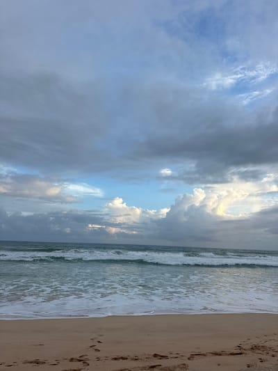 The image shows a beach scene with a dramatic sky. The left side of the image is dominated by a vast sky filled with a mix of dark, heavy clouds and bright, illuminated clouds that create a sense of depth and movement. The light filtering through the clouds suggests either early morning or late afternoon. The ocean is on the right, with gentle waves rolling onto the sandy shore. The water is a greenish-blue hue, and white foam from the waves creates a delicate line where it meets the sand. The sand itself is light brown and shows several footprints, indicating recent human presence. The overall mood is serene yet awe-inspiring, with the play of light and shadow in the sky adding a touch of drama to the natural beauty of the scene. The location is Koggala, Sri Lanka.