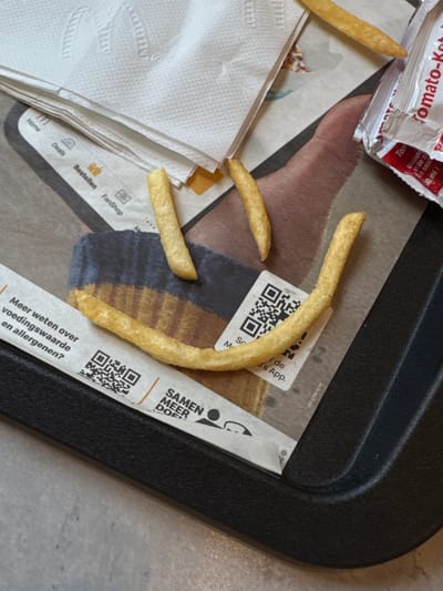 The image shows a close-up view of a McDonald's tray containing french fries, a ketchup packet, and napkins. The french fries are arranged in a smiley face formation on the tray. A ketchup packet, labeled "Tomato-Ketch," is visible on the right side of the image. Several napkins are stacked on the left side, one of which has the McDonald's logo imprinted on it. A QR code and text in Dutch are present on a paper strip at the bottom of the image. The background suggests an indoor setting, likely a McDonald's restaurant in Deventer, Netherlands, given the Dutch text. There are no people visible in the image.