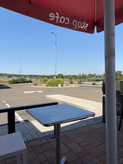 The image shows an outdoor seating area at a service station. In the foreground, a metal table with a light-colored stool is visible. Beyond the seating area, a paved road and a highway with traffic are depicted. The background features a clear blue sky, trees, and utility poles. The scene is set in Mogyoród, Hungary.
