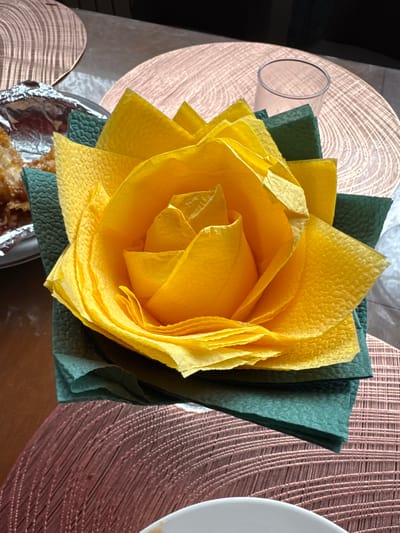 A close-up, eye-level shot shows a yellow paper napkin folded to resemble a rose, placed on top of a dark green napkin. The rose-shaped napkin has numerous layers of creased yellow paper, forming petals. The green napkin is folded into a square base and is positioned on a textured, round, copper-colored placemat. In the background, another placemat of the same design is visible, along with a clear glass and a corner of a foil-wrapped dish containing food. The scene appears to be set on a table. The location is not specified to be Chuguyevka, Russia.