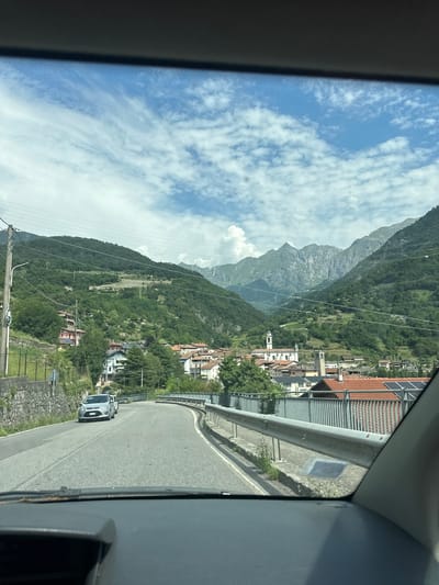 This media file shows a view from inside a vehicle, looking out at a scenic mountain landscape in Malegno, Italy. The primary subject is the setting: a winding paved road curving through a valley. On the left side of the road, a steep, forested mountainside rises dramatically, with rocky peaks visible in the distance. Nestled on the slopes of the mountain is a charming Italian village, characterized by traditional houses with red-tiled roofs and a prominent church steeple. Power lines crisscross the scene, and a guardrail runs along the right side of the road, bordering the drop-off.

In the foreground of the road, a silver SUV is driving away from the camera. The sky is bright blue with scattered white, fluffy clouds, suggesting a pleasant, sunny day. The time of day appears to be daytime, likely late morning or early afternoon, given the lighting.

The overall mood is one of travel and appreciation for natural beauty, with a hint of the quaintness of rural Italian life. There is no visible text in the image.