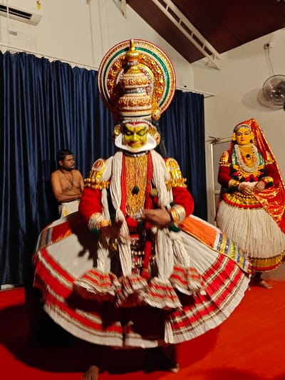 The image captures an indoor Kathakali performance at a dedicated venue in Kochi, Kerala, India. The central figure is a male performer in a vibrant Pacha vesham (green-faced character) costume, standing barefoot on a red carpeted stage. He wears elaborate green and red facial makeup, a prominent golden crown (Kireedam) with a circular green and gold halo, a red long-sleeved jacket adorned with gold, multiple gold necklaces, and a voluminous, layered white skirt featuring red and gold striped borders. His hands are positioned in a traditional mudra.
To the left of the central performer, a bare-chested man with dark hair and a beard, wearing a white dhoti and a string of beads, stands against dark blue curtains. He observes the primary performer. To the central performer's right, a second performer, likely portraying a female character (Stri vesham), stands in profile. This individual has yellow-orange facial makeup, a red and gold head covering, extensive gold jewelry, a red long-sleeved top, and a white pleated skirt with a colorful waistband.
The background consists of dark blue stage curtains, a white air conditioning unit mounted high on the left wall, and a dark brown sloped ceiling with white support beams. A black wall-mounted fan is visible on the upper right, and a white door frame is partially seen behind the rightmost performer. The scene is illuminated by interior lighting, casting a warm glow on the performers and stage.