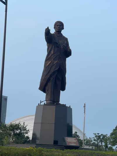 A statue of a man in a long coat and head covering stands tall against a clear, pale blue sky. The statue is dark bronze and the man's right arm is extended, with his index finger pointing forward. He holds a book in his left hand. The statue is mounted on a large, multi-sided dark gray pedestal. To the left of the statue, a tall, slender lamppost with lights is visible. In the background, a domed building with a reflective surface is partially obscured by trees. A tall, modern building with vertical lines is also visible to the far left. The foreground features a grassy slope with yellow flowers. A plaque is affixed to the front of the pedestal. The location is identified as Kosambi Barat, Indonesia.