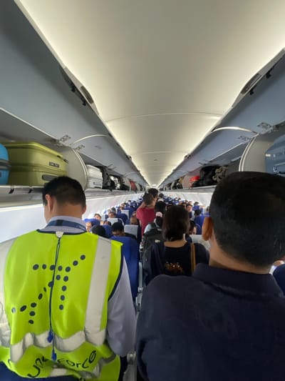 The image shows the interior of an airplane cabin. In the foreground, a person wearing a bright yellow safety vest with reflective stripes is facing away from the camera, looking down the aisle. The vest has a pattern of dark blue dots. Behind this person, another person is visible from the back, wearing a dark blue shirt.
The aisle is filled with passengers, most of whom are seated. Some passengers are looking towards the front or sides of the cabin. Luggage is stored in the overhead compartments, with several suitcases visible, including a lime green one. The cabin lighting is a bright, artificial white. The overall impression is that passengers are boarding or disembarking the aircraft, or preparing for take-off or landing. The specific location in Ahmedabad, India, cannot be determined from the image alone.