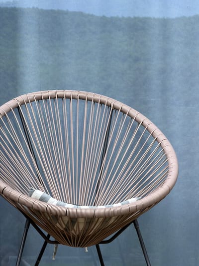 An overhead shot focuses on a light brown wicker chair set against a blue-gray mottled surface. The chair has a rounded back and is held up by a black metal frame. A checkered cushion sits inside. There are no people present, and the location of the city of Chkalov, Armenia, cannot be determined from the given visual context.