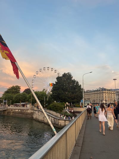 The image depicts a waterfront scene in Geneva, Switzerland, likely during the late afternoon or early evening, as indicated by the soft, warm lighting of the sky, which is a blend of pale blue and pastel orange hues. A large Ferris wheel, illuminated with white lights, stands prominently in the background, suggesting a fair or amusement park setting. The Ferris wheel has a colorful sign at its center.

In the foreground, a group of people are walking on a paved pathway next to a railing that overlooks a body of water. The people are dressed in casual summer attire. Some individuals are walking together, while others appear to be in smaller groups or alone. The water is calm, reflecting the ambient light and showing ripples on its surface. Beyond the pathway and railing, there are stone steps leading down to the water and more trees and buildings in the distance. One building has the text "ARAB BANK" and "RICHARD MILLE" visible on its facade. A flag, with red, black, and yellow stripes, is partially visible in the lower left corner of the image, attached to a metal pole. The overall atmosphere seems relaxed and pleasant, with people enjoying the waterfront.