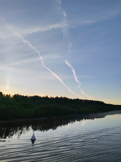 The image presents a tranquil landscape scene at what appears to be either sunrise or sunset over a body of water in Volokov Most, Russia. The sky dominates the upper half, displaying a gradient from a soft, warm yellow-orange hue near the horizon on the left, transitioning to a pale blue overhead towards the right. Several prominent, white aircraft contrails cut across the sky; one particularly long, wavy trail extends from the upper right, intersecting with another fainter trail closer to the upper left.
A dense, dark band of trees or forest lines the distant bank, casting a clear reflection onto the remarkably calm water below. The water occupies the lower half of the image, serving as a reflective surface for both the trees and the sky. In the lower left quadrant of the water, a white, conical buoy with a small dark top floats, creating subtle ripples around it and casting a dark reflection. More distinct, parallel wave patterns are visible across the water in the foreground, stretching from the right side towards the center.
The scene conveys a sense of peacefulness and natural beauty, with the atmospheric contrails adding a modern touch to the serene vista. There are no people visible, and no discernible text.