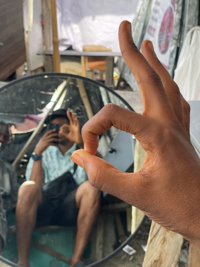 In Ojomu, Nigeria, a close-up shot focuses on a person's hand making the "okay" gesture in the foreground. Beyond the hand, a large circular mirror reflects a man in the background. This man is seated and holding a smartphone, seemingly taking a selfie. He is wearing a light blue, patterned short-sleeved shirt and dark shorts. A black crossbody bag is slung across his body. The mirror is positioned outdoors, with a background that appears to be an outdoor market or stall area, indicated by wooden structures, some covered by white tarps or fabric, and visible wares. The overall environment suggests a casual, everyday setting within the city.