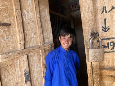 A young man with short black hair, wearing a blue collared shirt, smiles as he stands in a doorway. The doorway is made of wooden planks with writing on it in a language that is not English. The door is secured with a silver padlock. The door appears to be part of a structure that is not well-lit. The location is in Saranan, Pakistan, with coordinates of latitude: 30.532, longitude: 66.909.
