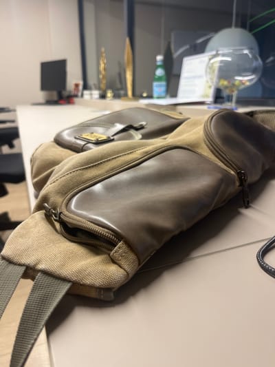 The image shows a close-up of a canvas and leather messenger bag, likely taken indoors. The bag is predominantly a light brown or tan canvas with darker brown leather accents and panels. It features multiple zippered compartments, with the zipper pulls clearly visible. A grey fabric strap is also visible, suggesting it's a shoulder or crossbody bag. The bag appears to be resting on a light-colored, smooth surface, possibly a desk or table.
In the background, which is out of focus, there are hints of a modern indoor setting. To the left, a glass sphere or orb is visible, containing what appears to be small, colorful objects or flowers. Below this, a partially visible bottle with a blue label, possibly a beverage, is present. Further to the left, there are reflections of lights and objects, including what looks like a computer monitor and some decorative items. The lighting suggests it might be artificial, possibly from overhead lights or a desk lamp, and the image has a slightly warm tone.
There is no discernible activity taking place, as the image focuses solely on the bag and its immediate surroundings. The overall mood is neutral and observational, simply documenting the object. No people are visible in the frame. The provided location context of Germasogeia Municipality, Cyprus, does not offer any direct visual cues within the image itself, but it suggests the general geographical area where the photo was taken.