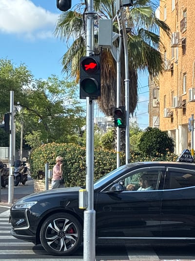 The image shows a street scene in Holon, Israel. A black car is stopped at a crosswalk with a red pedestrian signal. The traffic light for vehicles shows a green right turn arrow. A woman stands near the car, holding a phone. Several motorcycles are parked further down the street. A multi-story building is in the background with air conditioning units on the facade. Palm trees are visible.